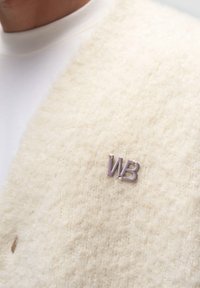Gilet in misto lana color avorio con una superficie morbida e testurizzata. Presenta una piccola spilla "WB" viola attaccata al tessuto.