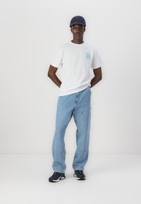 White cotton t-shirt with teal graphic, blue denim jeans, and black athletic shoes. Casual cap in dark blue. Standing with hands behind back.