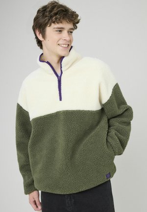 Pull en polaire avec un zip quart, présentant une partie supérieure crème et une section inférieure verte olive. Tissu texturé avec un zip violet et des détails de logo.