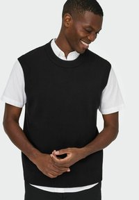 Schwarzer gestrickter ärmelloser Pullover mit Rundhalsausschnitt, über einem weißen kurzärmeligen Hemd getragen. Einfaches Design mit gerippten Abschlüssen.