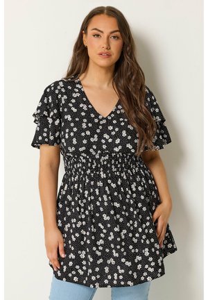 Mujer con pelo largo y castaño que lleva un vestido negro de manga corta con flores blancas de margarita, cintura fruncida y vaqueros azules, de pie junto a una pared blanca.