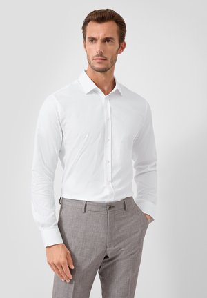 Mężczyzna w białej koszuli włożonej do jasnoszarych eleganckich spodni, stojący z jedną ręką w kieszeni na tle jednolitym.