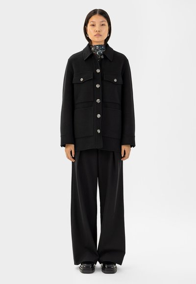 Schwarze Wollmischung Jacke mit Kragen, Fronttaschen und silbernen Knöpfen. Kombiniert mit weiten schwarzen Hosen und glänzenden schwarzen Schuhen.