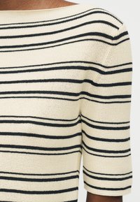 Κοντινό πλάνο ενός ατόμου που φοράει μπεζ πουλόβερ πλεκτό με οριζόντιες μαύρες ρίγες και μανίκια έως τον αγκώνα.