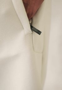 Pantaloni bej deschis cu o textură nervurată. Prezentă un fermoar negru cu un tab logo și un buzunar lateral vizibil în detaliu.