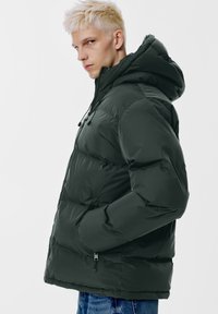Donkergroene gewatteerde jas met capuchon, voorzien van een rits aan de voorkant, een trekkoord en zijzakken. Gladde textuur met een vierkante pasvorm.