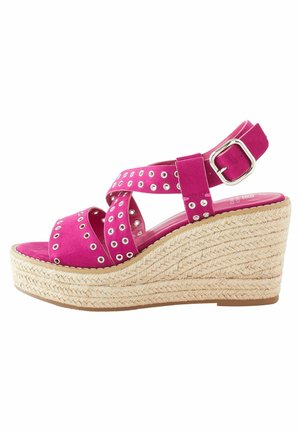 Rosa Wildleder-Wedge-Sandale mit gekreuzten Riemen, silbernen Ösenakzenten und einer geflochtenen Jute-Plattform. Verstellbarer Knöchelriemen mit Schnalle.