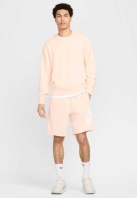 Sweatshirt rosa claro com punhos canelados, combinado com calções rosa claro que apresentam um logótipo branco da Nike. Meias brancas até ao tornozelo e sapatos claros.