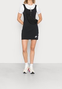 Svart bomullsklänning med rund halsringning, dragkedjestängning och en logotyp. Bärs över en vit t-shirt med svarta detaljer, ihop med färgglada sneakers.