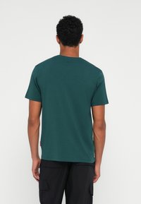 Tamsiai žalia medvilninė t-shirt su trumpomis rankovėmis ir apvalia iškirpte, turinti tiesų apatinį kraštą, rodoma iš nugaros. Nėra matomų raštų ar akcentų.