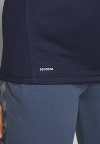Close-up van een persoon die een marineblauw 'BEECOOL' shirt en een blauwe broek met een grijze patroonstreep aan de zijkant draagt.