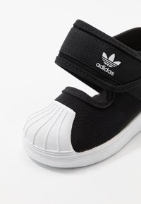 Svarta Adidas barnskor med vit gummiskalstå och krok-&-öglestängning mot vit bakgrund.