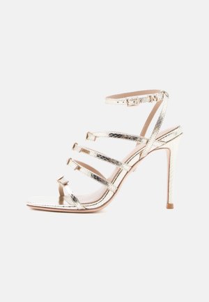 Sandalia de tacón alto metálica dorada con cuatro finas tiras cruzando el pie y una correa en el tobillo con hebilla.