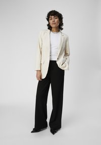 Blazer beige sur une chemise blanche, associé à un pantalon noir à jambes larges et des chaussures noires à bout pointu. Cheveux bouclés et bijoux minimalistes complètent le look.