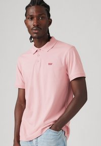 Polo en coton rose clair avec un col et trois boutons, présentant un petit logo rouge Levi's sur la poitrine. Manches courtes.