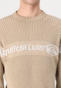 Beige gerippter Pullover aus Strickstoff. Mit einem weißen Logo über der Brust. Rundhalsausschnitt und lange Ärmel.
