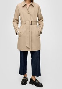 Osoba w beżowym płaszczu trench z paskiem, ciemnych, skróconych dżinsach i czarnych loafersach z złotym łańcuchowym detalem, stojąca na jednolitym tle.