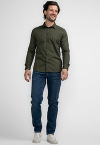 Camisa de botones verde con estampado, de manga larga, combinada con jeans azules y zapatillas blancas informales. Tela suave y diseño ajustado.