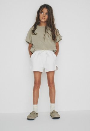 Dievča s dlhými hnedými vlasmi stojí s rukami v kapsách, oblečené v voľnom béžovom tričku, bielych šortkách, ponožkách a béžových šmykľavých topánkach na bielom pozadí.