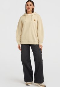Beżowy oversize'owy hoodie z kieszenią kangurzą i kapturem, z okrągłą naszywką logo na klatce piersiowej, w połączeniu z szerokimi czarnymi spodniami.