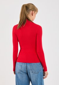 Rdeča rebrasta majica z dolgimi rokavi, ki ima pokrito vrat in prilega postavi, v kombinaciji z svetlo modrimi jeans hlačami z visokim pasom.