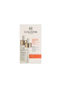 Bottiglia di plastica trasparente con conta-gocce, dettagli dorati, liquido bianco; confezione beige e arancione con logo del marchio e dettagli SPF 50. Indicatore di dimensione maxi.