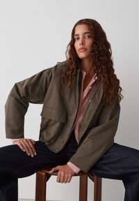 Olijfgroen cropped jack met een boxy fit, een voorzak en drukknoopsluiting, gelaagd over een lichtroze knopentrui en donkere jeans.