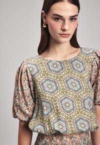 Blusa a maniche corte con motivi esagonali in verde, rosa e blu. Presenta dettagli arricciati sulle spalle e una morbida texture del tessuto.