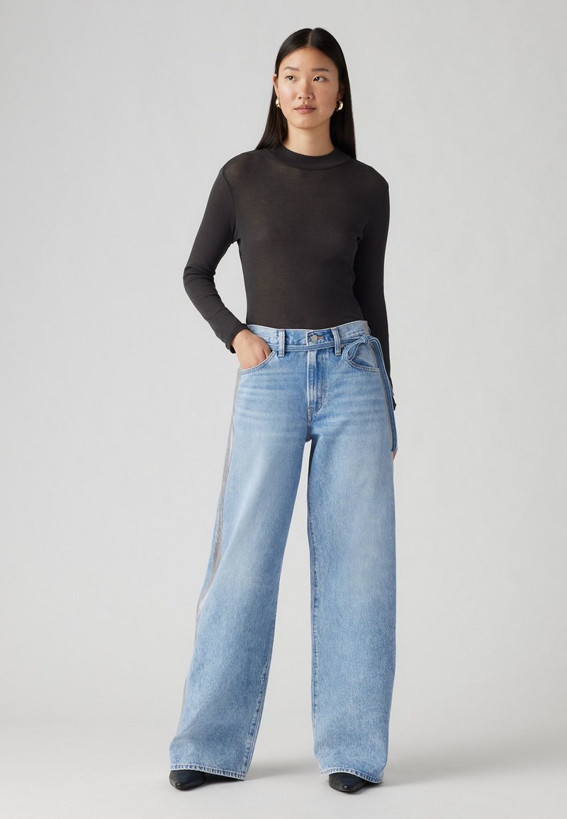 Maglietta nera a maniche lunghe abbinata a jeans a gamba larga blu chiaro con vita alta, tasche e dettaglio cintura annodata, su uno sfondo neutro.