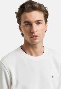 Weißes Sweatshirt mit geripptem Rundhalsausschnitt, weicher Textur und kleinem gesticktem Logo auf der linken Brust. Einfaches, sauberes Design.