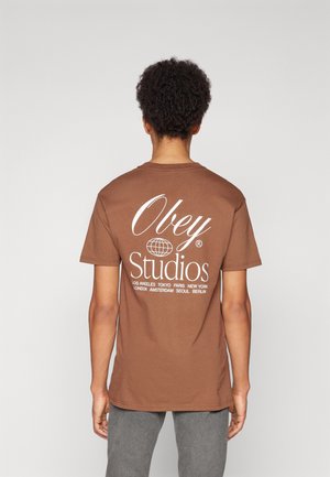Hnědý bavlněný tričko s bílou grafikou na zadní straně, která obsahuje text "Obey Studios" a názvy měst. Krátké rukávy, kulatý výstřih.