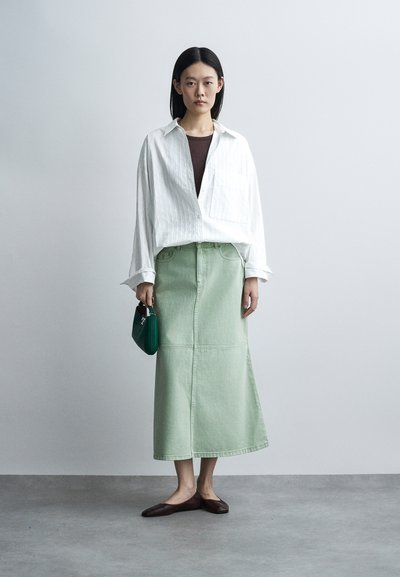 Femme debout portant une chemise blanche oversize, un haut marron, une longue jupe vert clair, tenant un petit sac à main vert, portant des ballerines marron sur un sol gris.