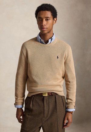 Jeune homme portant un pull beige par-dessus une chemise bleu clair et un pantalon marron à carreaux, debout devant un fond neutre uni.