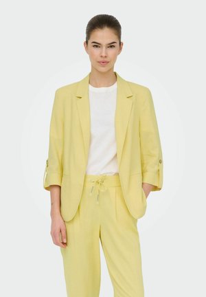 Femme debout, portant un blazer jaune clair aux manches retroussées et un pantalon à cordon assorti sur une chemise blanche, sur un fond uni.