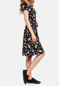 Kobieta ubrana w czarną sukienkę w kwiaty do kolan, z krótkimi rękawami i czarnymi sneakersami, stojąca w profilu na tle białym.