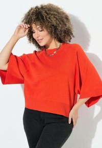 Roter Strickpullover mit kurzen, weiten Ärmeln und geripptem Saum; kombiniert mit schwarzen, figurbetonten Hosen. Glatte Textur, lässiges Design.