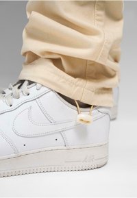 Białe skórzane sneakersy z szarymi sznurówkami i gumową podeszwą. Beżowe spodnie lekko zasłaniają buty, mające szczegół z zapięciem na sznurku. Tło jest szare.