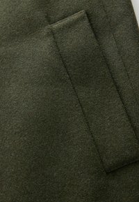 Dunkelgrüner Filzstoff mit einer rechteckigen aufgesetzten Tasche, glatte Textur mit leichten Oberflächenvariationen und einem matten Finish.