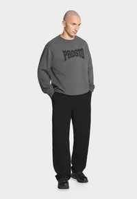 Szara bluza z nadrukiem "PROSTO", w połączeniu z czarnymi spodniami i czarnymi trampkami. Materiał wydaje się miękki i ma luźny krój.