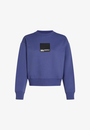 Sweatshirt roxo com um decote e punhos canelados. Apresenta um retângulo preto com texto em branco. Tecido em mistura de algodão, ajuste casual.