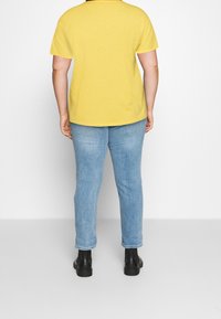 T-shirt giallo a maniche corte abbinato a jeans skinny blu chiaro e stivali neri alla caviglia, visto da dietro su uno sfondo bianco.