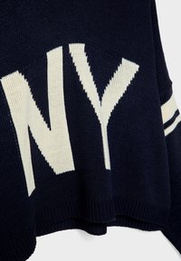 Pull en tricot bleu marine avec de grandes lettres blanches "NY" et des rayures blanches sur les manches, vu en gros plan sur la texture du tissu.