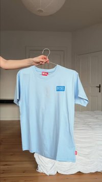 Ανοιχτό γαλάζιο T-shirt Diesel με λογότυπο στο στήθος και ετικέτα, κρεμασμένο σε σύρμα κρεμάστρας που κρατιέται από ένα χέρι σε ένα υπνοδωμάτιο με ξύλινο πάτωμα και λευκό κρεβάτι.