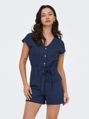 Femme portant une combinaison courte bleu marine à manches courtes avec des boutons et une ceinture nouée à la taille, debout devant un fond clair uni.
