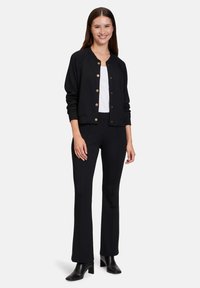 Cardigan noir à côtes avec des boutons dorés, porté sur un haut blanc, associé à un pantalon évasé noir et des bottines, sur fond blanc.