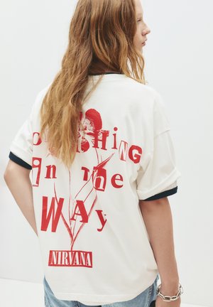 Fehér, oversized póló piros mintával. A feliratok így szólnak: "Crushing In the Way" és "NIRVANA". Kék akcentusokkal a ujjain.