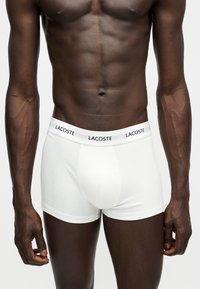 Homme musclé à la peau foncée portant un boxer blanc Lacoste avec logo sur la ceinture, debout sur un fond clair uni.