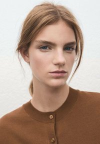 Cárdigan marrón de punto con un cuello redondo, que cuenta con una fila de botones, llevado sobre piel clara, con cabello liso de longitud media y ojos azules.