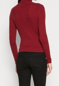 Roter, gerippter Rollkragenpullover mit figurbetonter Passform, der texturierte vertikale Linien und ein nahtloses Rücken-Design aufweist, getragen zu schwarzer Jeans.