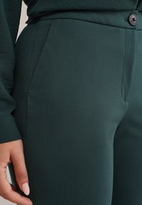 Tummanvihreät housut, joissa on sileä pinta, istuva malli, sivutaskut ja yksi napitus vyötäröllä.